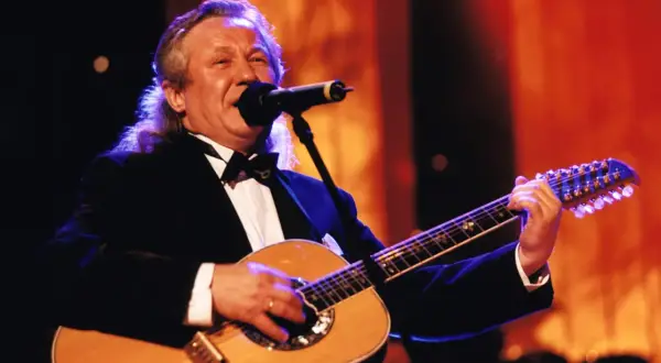 Artysta w czarnym smokingu gra na gitarze, śpiewając do mikrofonu. W tle pomarańczowe światła, jakby na scenie, gdzie kiedyś występowały siedem dziewcząt z Albatrosa.