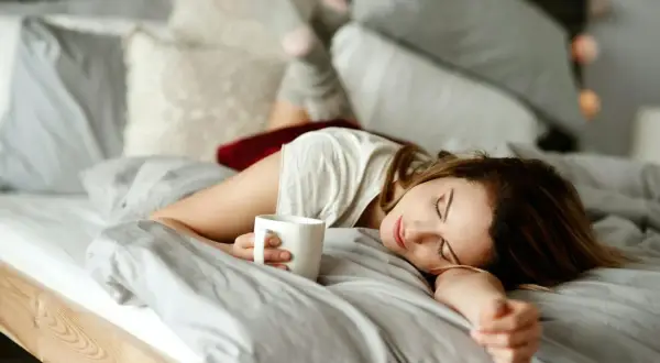 En kvinna sover i sängen med en kaffekopp i handen. Kaffe bra eller dåligt? Kanske en tupplur är bättre.