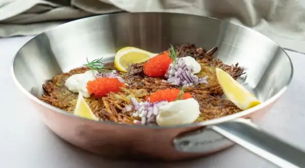 En gyllene råraka i stekpanna, toppad med gräddfil, rödlök, rom och citronklyfta. Ett klassiskt råraka recept som ser utsökt ut.
