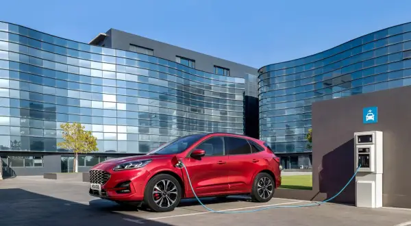 Czerwony Ford Kuga, nowoczesny kombi, ładuje się przy stacji ładowania przed futurystycznym budynkiem.