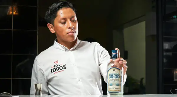 Młody mężczyzna w białej koszuli prezentuje butelkę tequili 30-30 Reserva Especial Blanco, podkreślając, z czego jest tequila – ze 100% niebieskiej agawy.