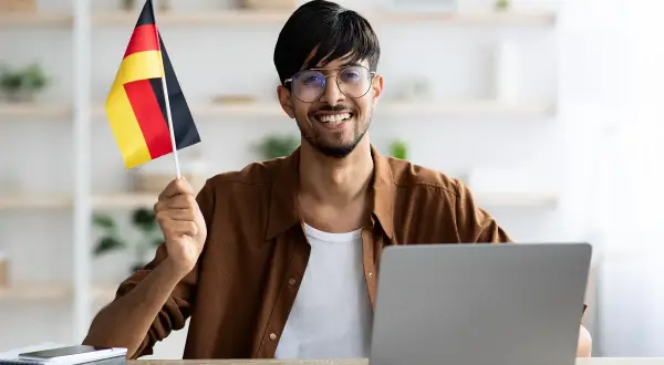 Uśmiechnięty mężczyzna z flagą Niemiec przy komputerze, planuje otwarcie konta w niemczech bez meldunku.