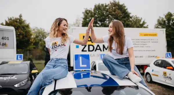 Dwie uśmiechnięte dziewczyny świętują sukces po egzaminie praktycznym kat B. Siedzą na dachu samochodu nauki jazdy, otoczone innymi pojazdami szkoleniowymi.