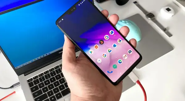 Dłoń trzyma smartfon podłączony kablem do laptopa, ułatwiając automatyczne przesyłanie zdjęć z telefonu na komputer.