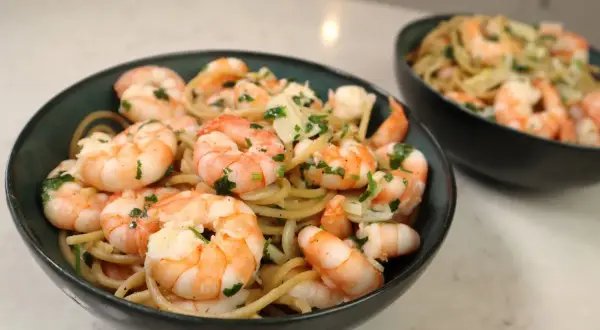 Pyszny makaron z krewetkami, posypany świeżą natką pietruszki i parmezanem, podany w ciemnych miseczkach.