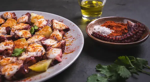 Un plat de poulpe à la gallega, garni de pommes de terre et d'épices, avec un petit bol de paprika et de sel, et un verre d'huile d'olive.