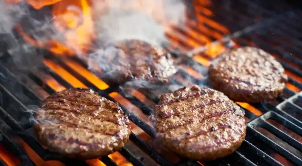 Soczyste burgery z grilla skwierczą na rozgrzanym ruszcie, otoczone płomieniami i dymem. Idealne na letnie popołudnie.