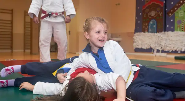 Niñas sonriendo mientras practican judo para niños. Un instructor de judo con cinturón rojo observa.