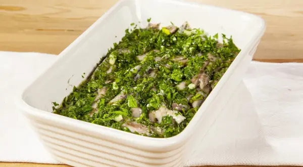 Acciughe al verde in una teglia bianca, condite con abbondante prezzemolo tritato e aglio.