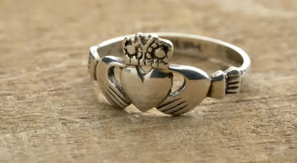 Anillo Claddagh de color oro blanco, con corazón, manos y corona, sobre madera.