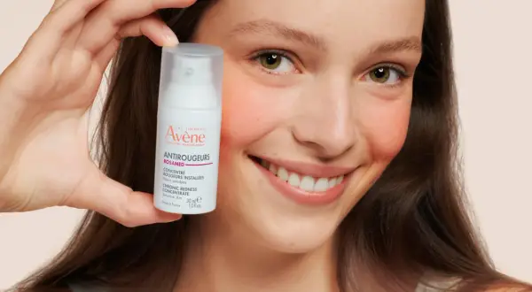 Ein lächelndes Gesicht hält eine Flasche Avène Antirougeurs. Ideal bei Rötungen, auch bei Neurodermitis.
