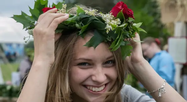 Uśmiechnięta dziewczyna w wianku z czerwonych róż i zielonych liści, jak na krakowskim festynie.