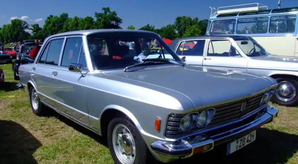 Srebrny fiat 130 z lat 70. prezentuje się elegancko na trawie, obok innych klasyków.