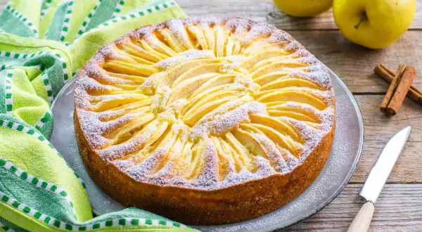 Torta di mele con yogurt, decorata con fettine di mela e zucchero a velo, su un piatto grigio, accanto a un tovagliolo verde e bastoncini di cannella.