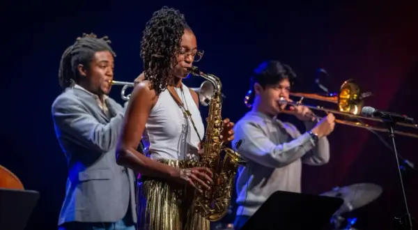 Zespół jazzowy gra na scenie. Kobieta gra na saksofonie, mężczyzna na trąbce, a drugi na puzonie. Inspiracja do tego, jak napisać tekst piosenki.