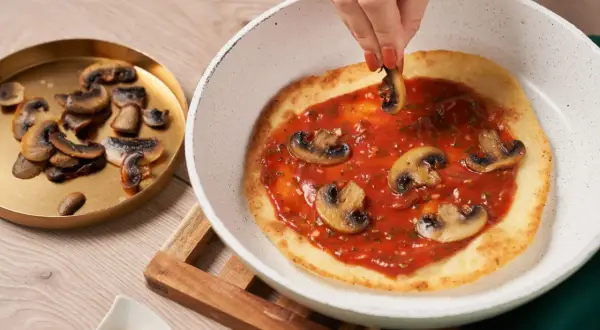 En hand lägger champinjoner på en pizza i stekpanna med tomatsås.