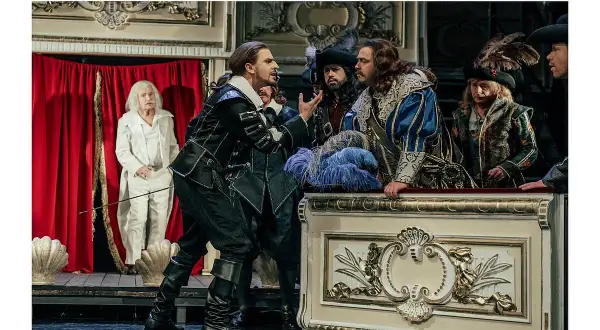Aktorzy w strojach z epoki na scenie, w tym mężczyzna w czarnym stroju, przypominający Cyrano de Bergerac, gestykuluje do towarzyszy.