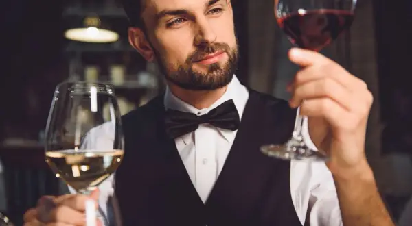 Elegancki mężczyzna w muszce ocenia dwa kieliszki wina. Pokazuje, jak trzymać kieliszek wina, by docenić jego barwę i aromat.