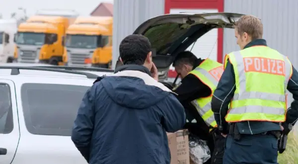Polizisten durchsuchen ein Auto. Ein Mann telefoniert, während die Beamten den Kofferraum inspizieren. Im Hintergrund stehen Lastwagen.