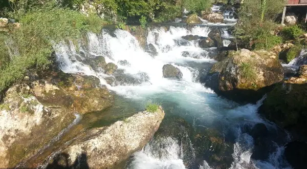 Vattenfall och klippor vid en flod nära Banja Luka. En liten röd stuga skymtar bland träden.