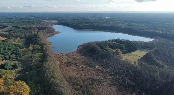 Malownicze jezioro wolickie otoczone lasami i polami. Spokojna tafla wody odbija błękitne niebo z chmurami.