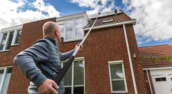 Hombre con pértiga extendida para limpiar tejas de una casa de ladrillo rojo bajo un cielo azul con nubes.