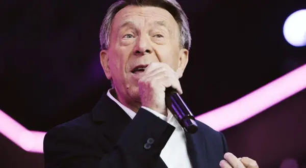 Wojciech Gąssowski śpiewa "Gdzie się podziały tamte prywatki". Artysta w czarnym garniturze i białej koszuli, z mikrofonem w dłoni, na tle fioletowych świateł.