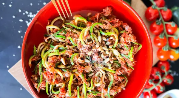 Ein leckeres Zoodles Rezept mit Hackfleischsauce, Pinienkernen und Parmesan in einer roten Schüssel. Daneben Kirschtomaten und Zucchini.