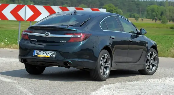 Ciemnozielony Opel Insignia 1.6 Turbo jedzie po drodze, za nim widać znaki ostrzegawcze i zielone pola.