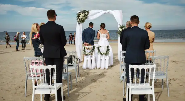 Piękny ślub na plaży. Para młoda stoi przed białym baldachimem, goście obserwują ceremonię.