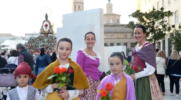 Niñas con trajes regionales y flores rojas, listas para las fiestas Zaragoza. Detrás, un gran tapiz floral y edificios históricos.