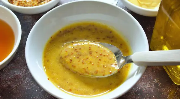 Cuchara con salsa miel y mostaza, lista para aderezar. Ingredientes en cuencos alrededor.