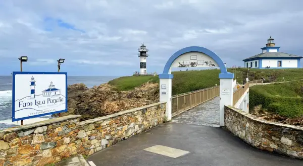 El faro de Ribadeo, con su icónico diseño blanco y negro, se alza sobre un acantilado verde. Un puente arqueado conduce a la isla.