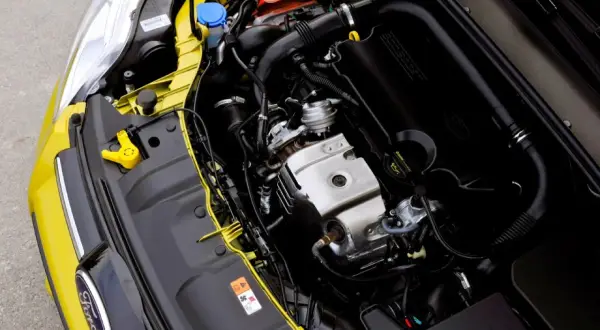 Silnik 1.0 Ecoboost w żółtym Fordzie. Widoczny jest czarny kolektor dolotowy z napisem "ECOBOOST" oraz metalowe elementy silnika.