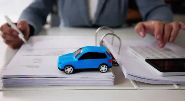 Osoba z długopisem i kalkulatorem analizuje dokumenty, obok stoi niebieski model auta. Zastanawia się, po jakim czasie można sprzedać auto.