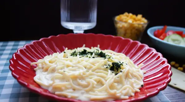 En krämig mac and cheese med persilja på en röd tallrik. Bredvid syns en sallad och ett glas vatten.