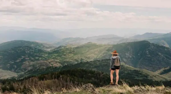 Una excursionista con sombrero admira las majestuosas montañas de Madrid, un paisaje verde y brumoso bajo un cielo nublado.
