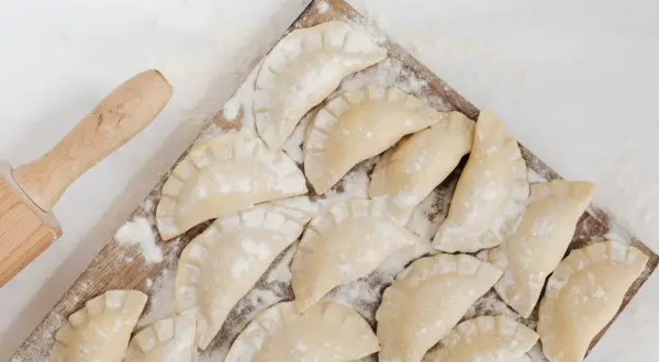 Surowe pierogi ruskie ułożone na desce, obok wałek do ciasta.