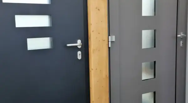 Dwie nowoczesne drzwi, jedna ciemnoszara z trzema prostokątnymi przeszkleniami, druga szara z trzema kwadratowymi, obok drewnianego słupka. Dowiedz się, jak wygłuszyć drzwi.