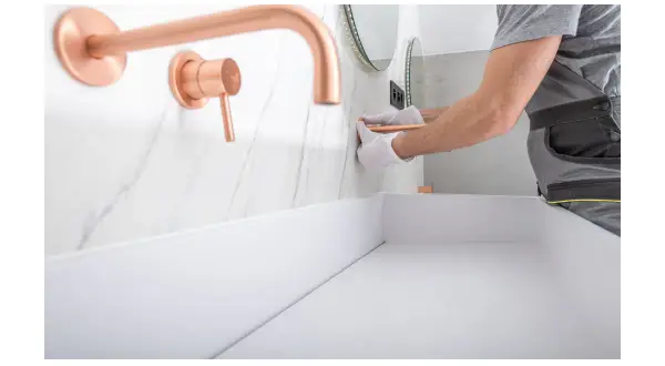 Instalator w białych rękawiczkach montuje miedzianą baterię podtynkową nad białym blatem.
