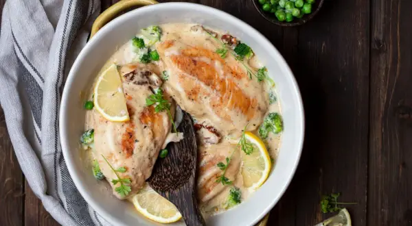Delizioso pollo al vino bianco in salsa cremosa con broccoli, piselli e fette di limone, guarnito con prezzemolo fresco.
