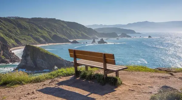 Un banco vacío invita a contemplar el mar azul desde el mirador de Coitelo, con acantilados verdes y rocas emergiendo del agua.