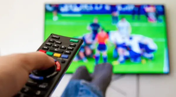 Dłoń trzymająca pilot do telewizora, na ekranie mecz piłki nożnej.