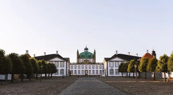 Frederiksborgs slott med sin gröna kupol reser sig majestätiskt mot en klar himmel. En stenlagd väg leder fram till den imponerande byggnaden, flankerad av träd.