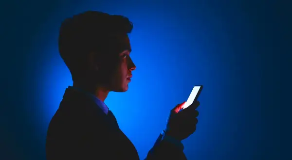 Mężczyzna w garniturze patrzy na jasny ekran telefonu. Dba o ochronę wzroku w telefonie, używając go w ciemności.