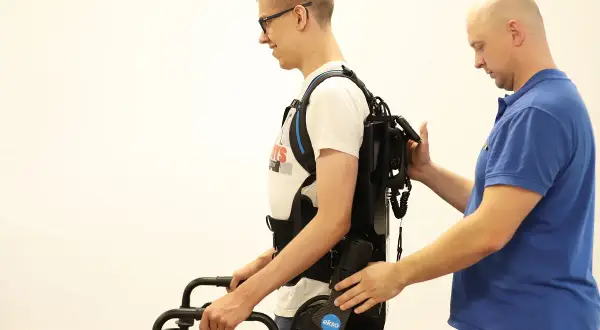 Mężczyzna w okularach ćwiczy chód w egzoszkielecie, wspierany przez terapeutę. Trójnóg rehabilitacyjny pomaga w pionizacji.