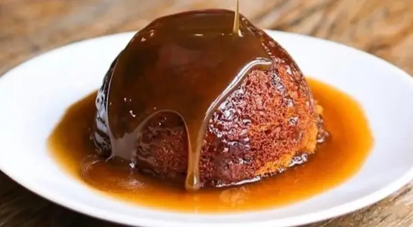 Un delizioso sticky toffee pudding, ricoperto da una cascata di salsa al caramello, servito su un piatto bianco.