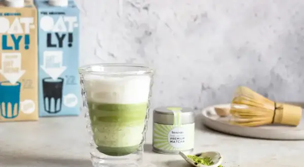 Warstwowy napój matcha z mlekiem owsianym, obok puszka z matchą i bambusowa miotełka. Idealny matcha przepis.