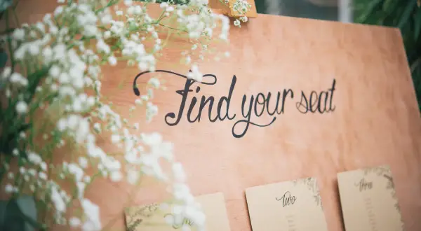 Panneau en bois avec l'écriture "Find your seat" et des fleurs. Des cartes de placement sont fixées en dessous.