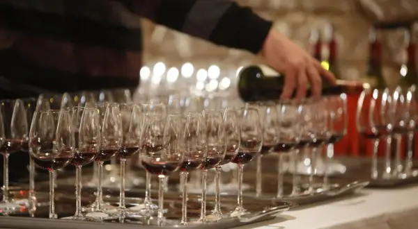 Rzędy kieliszków z czerwonym winem, osoba nalewająca wino z butelki.
